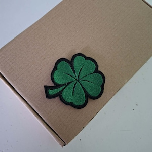 May include: A four-leaf clover patch featuring vibrant green embroidery with a distinct black border. The clover design includes four rounded heart-shaped leaves and a small stem extending to the left, all rendered in a dense, textured thread.