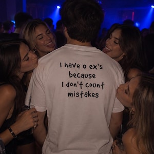 Può includere: Una t-shirt bianca a maniche corte con testo nero sul retro che recita: I have 0 ex's because I don't count mistakes. La maglia presenta un girocollo e una vestibilità standard, con il messaggio stampato chiaramente sulla parte alta della schiena.