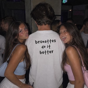 Puede incluir: Una camiseta blanca de manga corta con el texto negro "brunettes do it better" impreso en la parte posterior. La prenda tiene cuello redondo y un corte estándar, mostrada desde atrás con el texto claramente visible en el centro.
