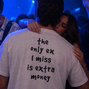 Może przedstawiać: Biały t-shirt z krótkim rękawem z czarnym napisem na plecach o treści, the only ex I miss is extra money. Koszulka jest noszona w słabo oświetlonym otoczeniu z niebieskim światłem, ukazując tył odzieży oraz nadrukowaną wiadomość na materiale.