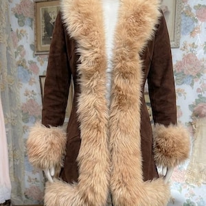May include: A brown suede coat featuring a long, thick beige faux fur trim along the front opening, collar, and sleeve cuffs. The coat is worn over blue denim jeans. The garment has a fitted silhouette and reaches mid-thigh length.