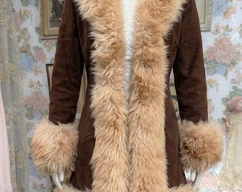 May include: A brown suede coat featuring a long, thick beige faux fur trim along the front opening, collar, and sleeve cuffs. The coat is worn over blue denim jeans. The garment has a fitted silhouette and reaches mid-thigh length.