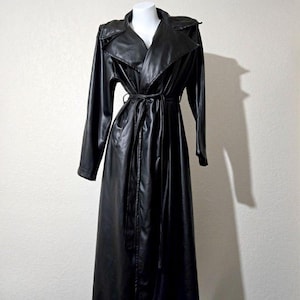 Puede incluir: Una gabardina larga de color negro con un acabado liso y reflectante. El abrigo presenta solapas anchas, mangas largas y un cinturón de lazo a juego en la cintura. Se muestra sobre un maniquí, destacando un cuello estructurado y falda larga.