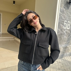 May include: A black zip-up jacket featuring a classic collar, long sleeves with buttoned cuffs, and two large front patch pockets with flaps. The jacket is constructed from a solid black fabric and is paired with blue denim jeans and amber-tinted sunglasses.