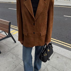 May include: A brown suede blazer with notched lapels and two front buttons, worn over a black turtleneck sweater. The outfit includes blue denim jeans, black pointed-toe shoes, and a black leather handbag with gold-tone hardware accents.