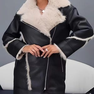 May include: A black jacket with a smooth finish, featuring a large cream-coloured faux fur collar and matching faux fur trim along the cuffs and hem. The jacket includes a silver-toned metal zip closure that runs diagonally across the front.