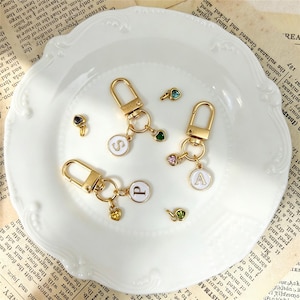 Op de afbeelding: Drie goudkleurige metalen sleutelhangers met ronde witte emaille bedels met de letters S, P en A. Elke sleutelhanger bevat een kleine hartvormige bedel met een gekleurde edelsteen. Drie extra losse hartvormige edelsteenbedels zijn inbegrepen.