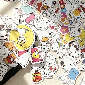 May include: A collection of die-cut vinyl stickers featuring the cartoon character Snoopy in various poses and activities. The stickers display vibrant colors including yellow, pink, red, and blue, with Snoopy interacting with items like gifts, books, and flowers.