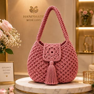 May include: A dusty rose crochet handbag featuring a textured star-stitch pattern, a single shoulder strap, and a central flap closure adorned with a circular mandala-style motif and a long, matching yarn tassel hanging from the center.