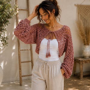 May include: A dusty rose crochet bolero featuring long, loose sleeves, an open front design with a tie closure, and decorative tassels. The garment is crafted with an intricate, open-knit pattern and is worn over a simple white tank top.