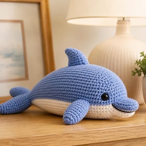 May include: A crocheted dolphin toy featuring a light blue upper body and a white underbelly. The plush figure has a prominent dorsal fin, two side fins, a tail, and small black plastic eyes. The texture is created with tight yarn stitches.
