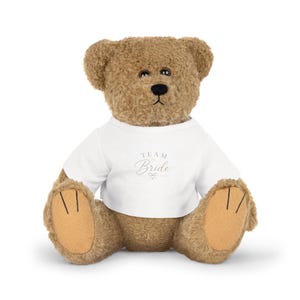 Puede incluir: Un oso de peluche marrón que lleva una camiseta blanca. La camiseta presenta el texto "TEAM BRIDE" impreso en una fuente cursiva gris, acompañado de un pequeño icono de corazón y un gráfico de lazo decorativo centrado en el pecho.