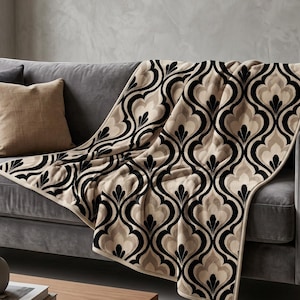 May include: A rectangular throw blanket featuring a repeating damask-inspired pattern in black, cream, and beige. The fabric has a soft, plush texture with finished edges, draped over a gray sofa alongside a solid tan square throw pillow.