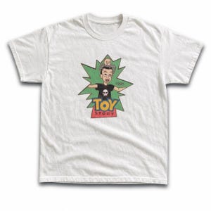 May include: A white short-sleeved t-shirt featuring a graphic of a cartoon character with a skull on their shirt, positioned in front of a green starburst shape. The text TOY STORY is printed below the character, with the year 1995 visible nearby.