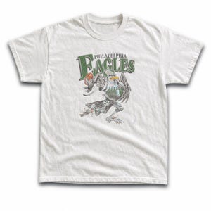 May include: A white short-sleeved t-shirt featuring a graphic of a cartoon eagle wearing a football helmet and jersey while running with a football. The text Philadelphia Eagles is printed in green above the eagle illustration in a bold font.