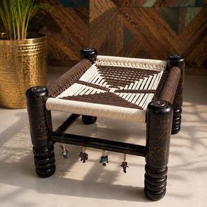 May include: A low stool with four dark brown, turned wooden legs featuring gold-toned painted details. The seat is woven with thick, cream and dark brown cord in a geometric pattern. Four small, colourful, dangling charms hang from the front rail.