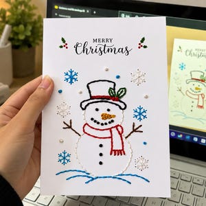 Puede incluir: Una tarjeta de felicitación blanca con un muñeco de nieve bordado a mano que lleva un sombrero de copa negro con acebo verde y una bufanda roja. El diseño incluye copos de nieve bordados en azul y blanco, pequeñas cuentas y el texto Merry Christmas.