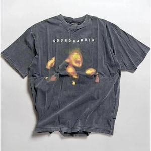May include: A charcoal gray short-sleeve t-shirt featuring the text SOUNDGARDEN in white capital letters above a blurry, abstract graphic of a face in shades of yellow, orange, and green. The fabric has a faded, washed-out appearance throughout.