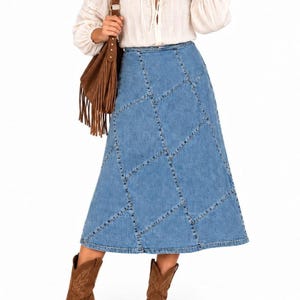 May include: A long blue denim skirt featuring a patchwork design with visible stitched seams, paired with a white long-sleeved blouse, a brown suede fringe shoulder bag, and brown leather boots with decorative stitching on the front.