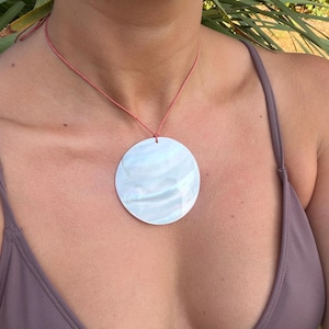 May include: A large, circular, iridescent mother-of-pearl pendant suspended from a thin, salmon-coloured cord. The pendant features a smooth, reflective surface with subtle white and pearlescent tones, hanging centred on a simple cord necklace.