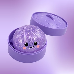 May include: A purple dumpling-shaped toy with a glittery texture, featuring a smiling face with black eyes and pink cheeks, resting inside a matching purple plastic steamer basket with a woven lid design and a small handle on top.