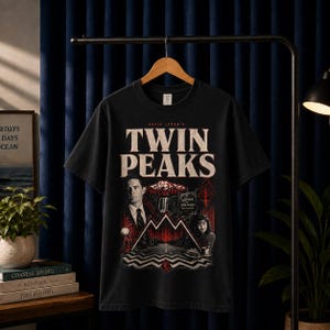Può includere: Una t-shirt nera a maniche corte con una stampa grafica recante il testo 'David Lynch's Twin Peaks'. Il design include i ritratti di due personaggi, un paesaggio montano, una cascata, una strada e un motivo a chevron rosso e nero.