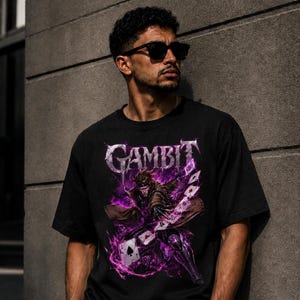 May include: A black short-sleeved t-shirt featuring a graphic print of the character Gambit. The design includes the text GAMBIT in a metallic font above the character, who is surrounded by purple energy effects and floating playing cards.