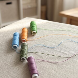 May include: Five spools of colored thread arranged in a staggered formation. The spools are light blue, orange, bright green, olive green, and purple. Each spool has a white plastic core and a single strand of matching thread extending outward.