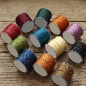 May include: A collection of twelve small spools of waxed thread in various colors including dark red, forest green, orange, light purple, olive green, teal, cream, black, navy blue, bright orange, and brown, each with a white circular label.