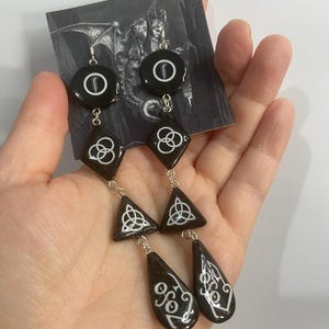 May include: A pair of long dangle earrings featuring black resin components in circular, diamond, triangular, and teardrop shapes. Each piece is decorated with white symbols, including a fern, triquetra, and stylised lettering, connected by silver-toned metal links.