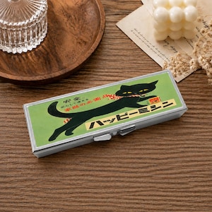 May include: A rectangular metal case with a hinged lid featuring a green background and a black cat illustration. The design includes Japanese text and a small red and white checkered pattern. The case has a silver-toned metallic frame and a latch.