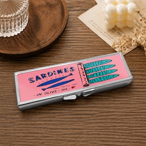May include: A rectangular silver metal case featuring a pink lid with a graphic illustration of four teal sardines in an open tin, two dark blue fish, and the text SARDINES IN OLIVE OIL with a small red heart. The case has a hinged lid.