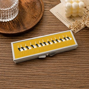 May include: A rectangular silver metal case featuring a mustard yellow top panel decorated with a repeating pattern of white and black cranes in profile. The case has a hinged lid with a metallic latch mechanism on the front edge for secure closure.