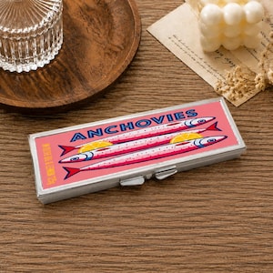 May include: A rectangular metal case with a pink background featuring a graphic illustration of three anchovies and two lemon slices. The text ANCHOVIES is printed in blue, and the text IN OLIVE OIL & LEMON 125g is printed in yellow.