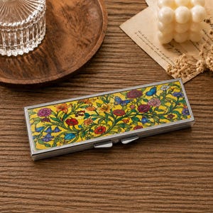 Peut inclure: Un étui métallique rectangulaire présentant un fond jaune vif orné d'un motif botanique détaillé composé de fleurs colorées, de vignes vertes et de petits papillons bleus. L'étui possède un cadre argenté poli et une fermeture à charnière.
