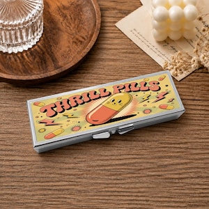 May include: A rectangular metal pill case featuring a graphic design with the text THRILL PILLS in a retro font. The illustration includes a smiling anthropomorphic capsule character, lightning bolts, and small starbursts on a light yellow background.