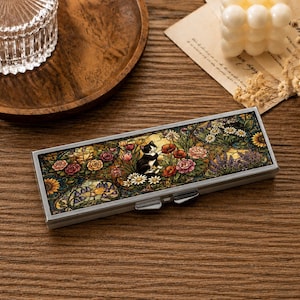 May include: A rectangular metal case featuring a stained glass style illustration of a black and white cat sitting among colourful flowers including red poppies, yellow sunflowers, and pink roses, with a silver metallic frame and a push-button latch.
