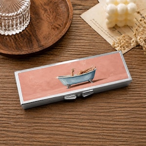 May include: A rectangular silver metal case featuring a printed illustration of a person relaxing in a light blue clawfoot bathtub against a solid dusty pink background. The case has a hinged lid with a secure latch mechanism on the front edge.