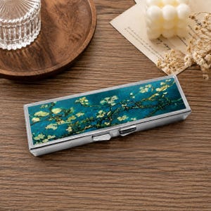 Puede incluir: Un estuche rectangular de metal con marco plateado y tapa con bisagras. La superficie superior presenta una reproducción impresa de una pintura que muestra flores de almendro blancas sobre un fondo azul verdoso profundo.