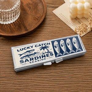 Może przedstawiać: Prostokątne metalowe pudełko z otwieraną pokrywką, ozdobione kremowo-granatowym wzorem graficznym. Nadruk zawiera napis LUCKY CATCH SARDINES SEASIDE, ilustracje dwóch pływających ryb, palmę oraz cztery pionowe ikony ryb.