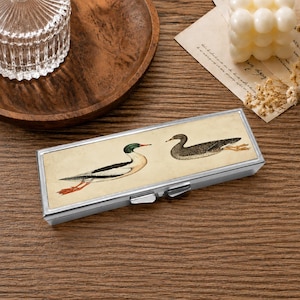 May include: A rectangular metal case with a hinged lid featuring a vintage-style illustration of two ducks on a cream background. One duck is depicted in profile with green, black, and white plumage, while the other is shown in flight with brown tones.