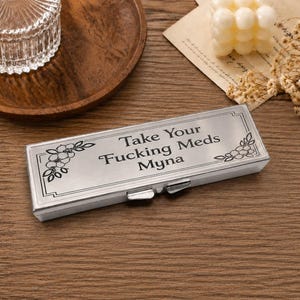May include: A rectangular silver-toned metal pill case with a hinged lid. The top surface is engraved with the text "Take Your FUCKING MEDS Myna" in black lettering, framed by decorative floral line art designs on the left and right corners.