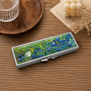 May include: A rectangular metal case featuring a vibrant reproduction of a floral painting on its lid. The artwork displays a dense field of blue irises and yellow flowers with green foliage. The case has a silver-toned metallic frame and a latch.