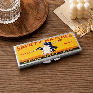 Pode incluir: Uma caixa metálica retangular com tampa articulada apresentando um gráfico de estilo vintage de um pinguim segurando um fósforo. O rótulo amarelo contém o texto SAFETY MATCHES, TRADE MARK e NIPPON MATCH CO., LTD. em letras pretas.