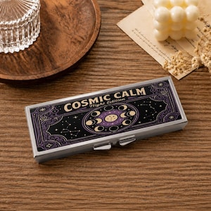 Pode incluir: Uma caixa metálica retangular com moldura prateada, apresentando um design gráfico preto e roxo na tampa. O texto "COSMIC CALM Night Edition" está impresso em branco, acompanhado por ilustrações de fases lunares e constelações.
