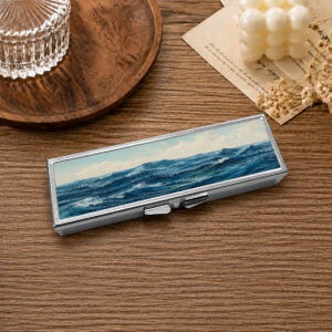 May include: A rectangular metal case featuring a printed image of a turbulent ocean with deep blue waves and white foam under a cloudy sky. The case has a silver-toned frame with a hinged lid and a small latch mechanism on the front edge.