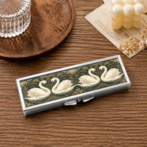 May include: A rectangular metal case featuring a decorative top panel with a dark green background and a repeating pattern of white swans swimming among gold-toned foliage. The case has a silver-colored metallic frame and a hinged lid with two latches.