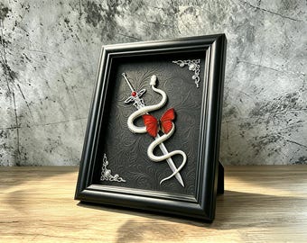 May include: A black framed wall art piece featuring a silver sword entwined with a white snake and a vibrant red butterfly. The background is a black textured surface with intricate scrollwork patterns and silver decorative corner accents.
