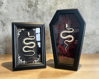 May include: Two framed displays featuring snake skeletons. One is a rectangular black frame with a white snake skeleton on a black background. The other is a coffin-shaped black frame containing a white snake skeleton over a dark red rose pattern.