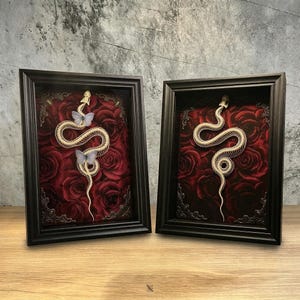 May include: Two rectangular black frames containing white snake skeletons displayed against a deep red rose background. The left frame features two light blue butterflies perched on the skeleton, while the right frame shows only the snake skeleton.